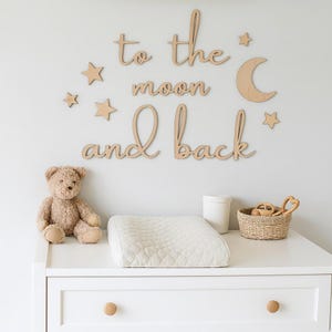 Może przedstawiać: Drewniane dekoracje ścienne do pokoju dziecięcego z napisem "to the moon and back", gwiazdami i księżycem. Miś, przewijak i kosz z zabawkami stoją na białej komodzie.