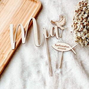 May include: Wooden cake topper with the name "Mia" in cursive script, a dove with an olive branch, and a fish with the text "Confirmation 22.05.2022".
