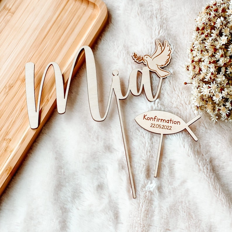 May include: Wooden cake topper with the name "Mia" in cursive script, a dove with an olive branch, and a fish with the text "Confirmation 22.05.2022".