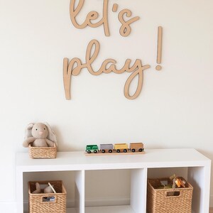 May include: A white storage unit with three cubbies, holding woven baskets and a plush bunny. A wooden train set sits on top, and a large wooden sign that says "let's play!" is mounted on the wall.