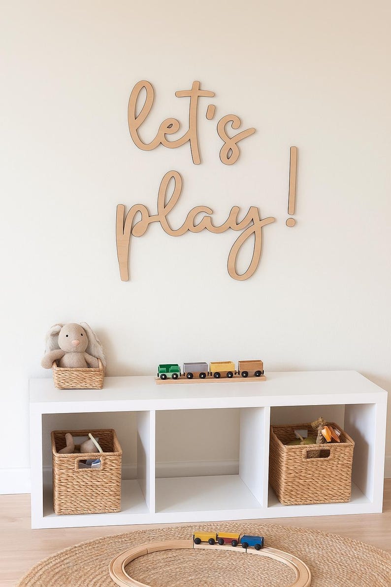 May include: A white storage unit with three cubbies, holding woven baskets and a plush bunny. A wooden train set sits on top, and a large wooden sign that says "let's play!" is mounted on the wall.