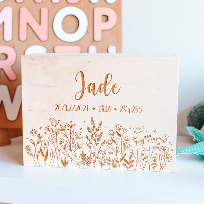 May include: A light-colored wooden box with the name "Jade" engraved on the top. The box features an engraved floral design along the bottom edge and birth details: 20/12/2021, 1h14, and 2kg 285.