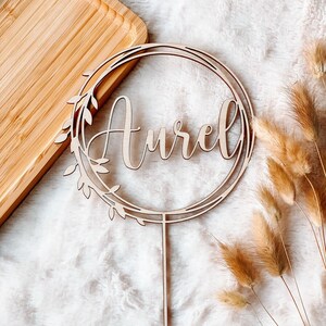 May include: A wooden cake topper with a circle design and the name "Aurel" written in cursive. The topper is on a white fuzzy surface with dried grass.