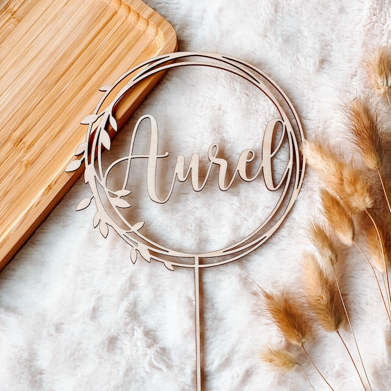 May include: A wooden cake topper with a circle design and the name "Aurel" written in cursive. The topper is on a white fuzzy surface with dried grass.