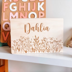 May include: A light-colored wooden sign engraved with the name "Dahlia" in a cursive font. The bottom of the sign features an engraved floral design with flowers, leaves, and a butterfly.