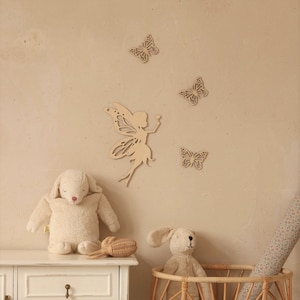 Pu&ograve; includere: Decorazioni murali in legno a forma di fata e farfalle. La fata tiene in mano un piccolo fiore. Tre farfalle sono sparse intorno alla fata. Un agnello di peluche e un coniglietto di peluche sono su una cassettiera bianca e in un cestino di vimini.