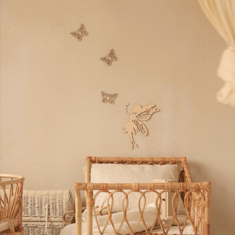Pu&ograve; includere: Decorazioni murali in legno raffiguranti una fata e farfalle. La fata ha le braccia tese e ali dettagliate. Tre farfalle sono sopra la fata. L'arredamento &egrave; marrone chiaro e montato su una parete beige. Una struttura letto in vimini &egrave; in primo piano.