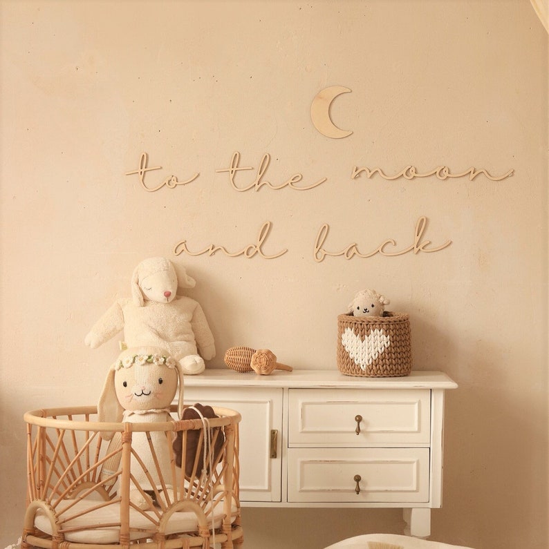 May include: Wooden wall decor with the phrase "to the moon and back" in cursive script. A crescent moon cutout is above the phrase. A white wicker bassinet with a stuffed bunny inside is in the foreground. A white dresser with two drawers is in the background.