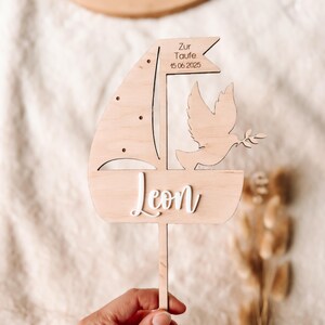 Può includere: Cake topper in legno con un design a forma di barca a vela e una colomba con un ramoscello d'ulivo. Il nome "Leon" è in corsivo bianco. La bandiera recita "Zur Taufe 15.06.2025".