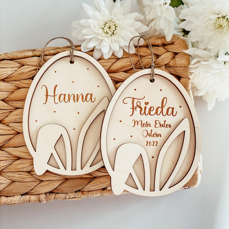 May include: Two wooden Easter egg decorations with bunny ear cut-outs. One ornament is personalised with the name "Hanna" and the other with the name "Frieda" and the text "Mein Erstes Ostern 2022".