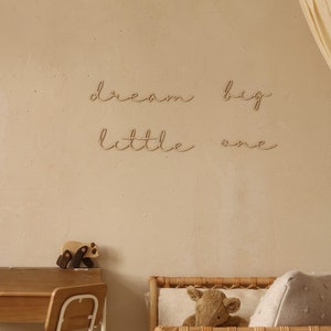 May include: Wooden wall decor with the words "dream big little one" in a cursive font. The decor is hanging on a light beige wall in a nursery setting.