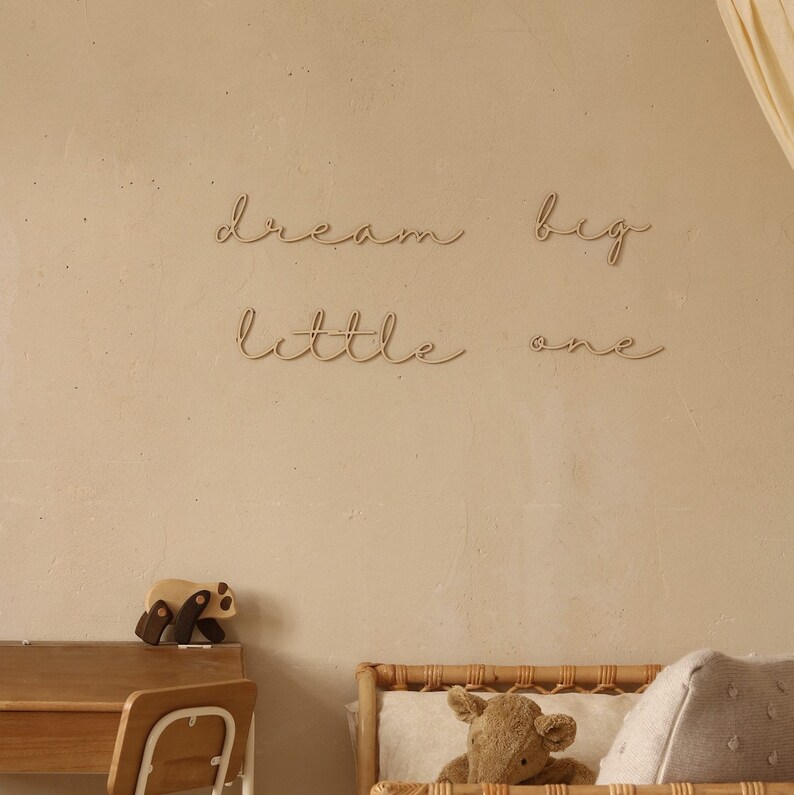 May include: Wooden wall decor with the words "dream big little one" in a cursive font. The decor is hanging on a light beige wall in a nursery setting.