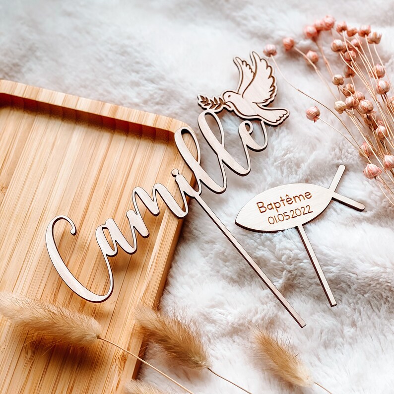 May include: Wooden cake topper with the name "Camille" in cursive script, a dove with an olive branch, and a fish with the text "Bapt&ecirc;me 01.05.2022".