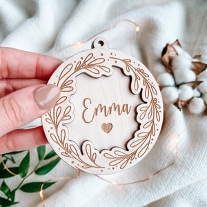 Op de afbeelding: Ronde houten ornament met de naam "Emma" in sierlijke letters, omringd door een bladerkrans. Een klein hartje is onder de naam gegraveerd. Het ornament heeft een gat aan de bovenkant om op te hangen.