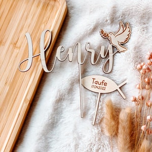 May include: Wooden cake topper with the name "Henry" in cursive script, a dove with an olive branch, and a fish with the text "Taufe 17.04.2022".