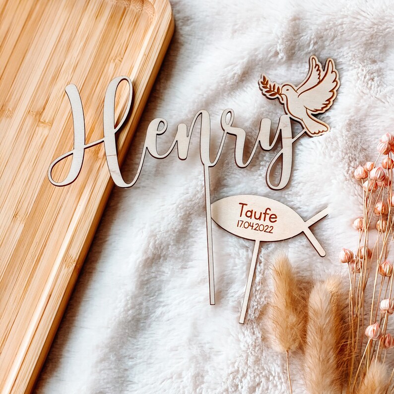May include: Wooden cake topper with the name "Henry" in cursive script, a dove with an olive branch, and a fish with the text "Taufe 17.04.2022".