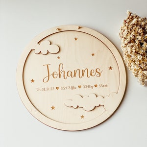 May include: A round wooden plaque with a cloud and star design. The plaque is engraved with the name "Johannes", the date "25.01.2022", the time "05:03 Uhr", the weight "3340g", and the length "55cm".