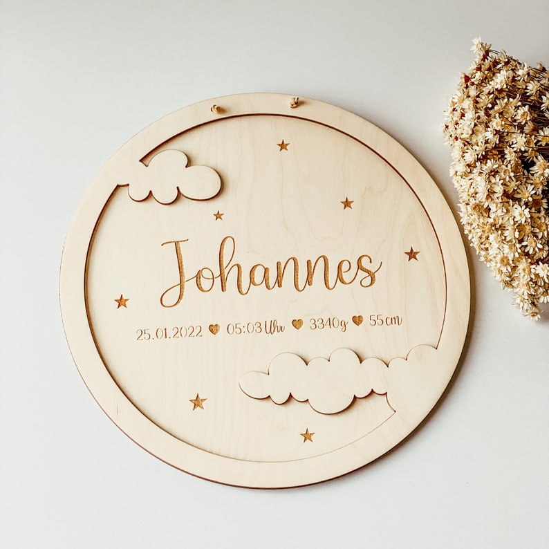 May include: A round wooden plaque with a cloud and star design. The plaque is engraved with the name "Johannes", the date "25.01.2022", the time "05:03 Uhr", the weight "3340g", and the length "55cm".