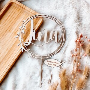 May include: Wooden cake topper with the name "Lina" in a circle with leaves around it. The topper also has a small wooden sign with the text "Konfirmation 5. Juni 2022" and a fish symbol.
