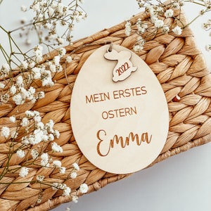 May include: A wooden Easter egg ornament with the text "Mein erstes Ostern Emma" and the year "2022" on a bunny cutout. The ornament is sitting on a woven basket.