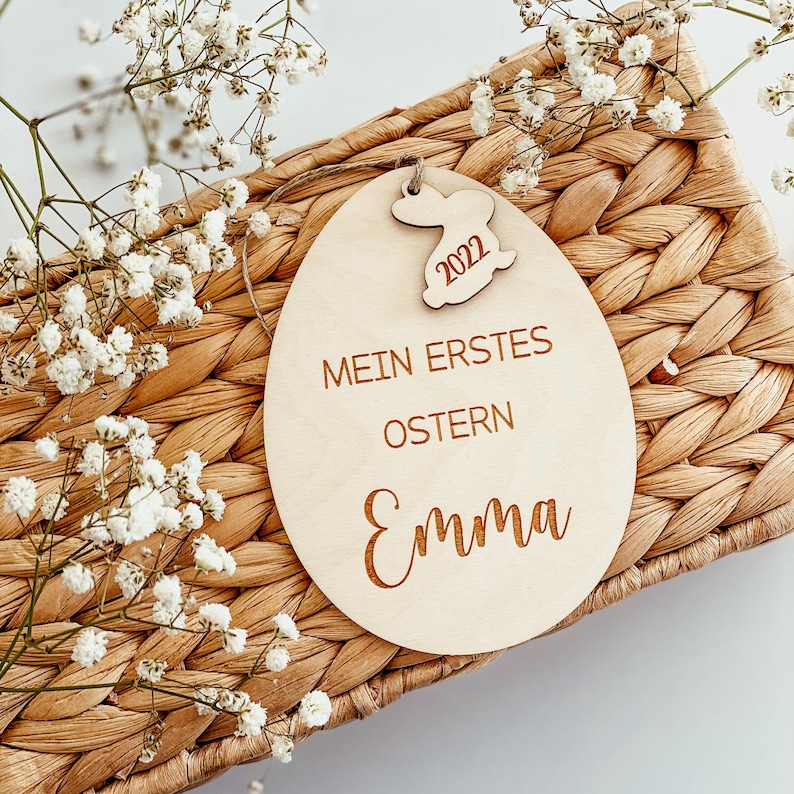 May include: A wooden Easter egg ornament with the text "Mein erstes Ostern Emma" and the year "2022" on a bunny cutout. The ornament is sitting on a woven basket.