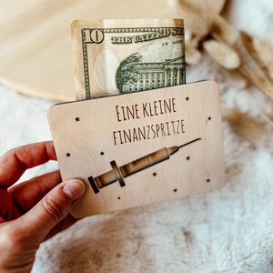 May include: A wooden card with the text "EINE KLEINE FINANZSPRITZE" and a syringe illustration. A ten-dollar bill is inserted into the card. The card has small star designs around the text and illustration.