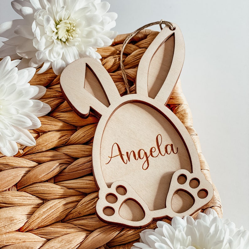 Pu&ograve; includere: Un porta uova di Pasqua in legno a forma di coniglio con il nome "Angela" inciso su di esso. Il porta uova &egrave; seduto su un cesto di vimini intrecciato.