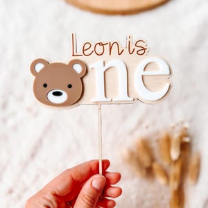Puede incluir: Un adorno para tarta de madera con el texto "Leon's one" y una cara de oso marr&oacute;n. La palabra "one" es blanca, y el oso tiene una cara marr&oacute;n claro con nariz y ojos negros. El adorno se sujeta con un palito de madera.