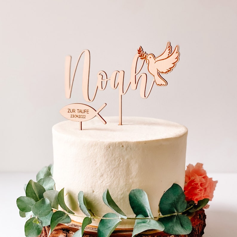 May include: Wooden cake topper with the name Noah, a dove with an olive branch, and a fish with the text "Zur Taufe 23.04.2022".