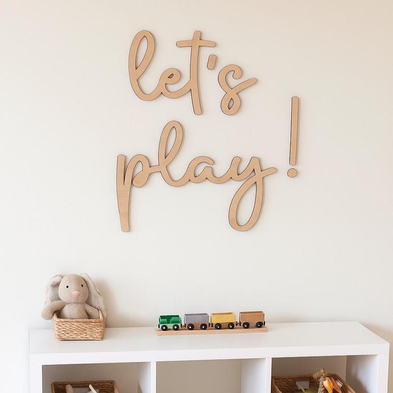 May include: Wooden wall art spelling "let's play!" in a cursive font. Below, a white shelf displays a stuffed bunny in a wicker basket and a wooden train set with colorful cars. The wall is a neutral color.