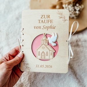 May include: A rectangular wooden plaque featuring the engraved text ZUR TAUFE von Sophie and 31.05.2026. The center displays a circular cutout with a pink background, an engraved church illustration, and a white dove cutout, bound with white ribbon.