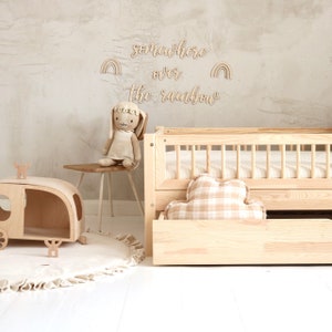 K&ouml;nnte beinhalten: Ein Kinderbett aus Holz mit einem wei&szlig;-braunen karierten Kissen und einem h&ouml;lzernen Spielzeug-Wohnwagen. Die Wand hinter dem Bett hat ein Holzschild mit der Aufschrift "somewhere over the rainbow" mit zwei Regenbogen-Ausschnitten.