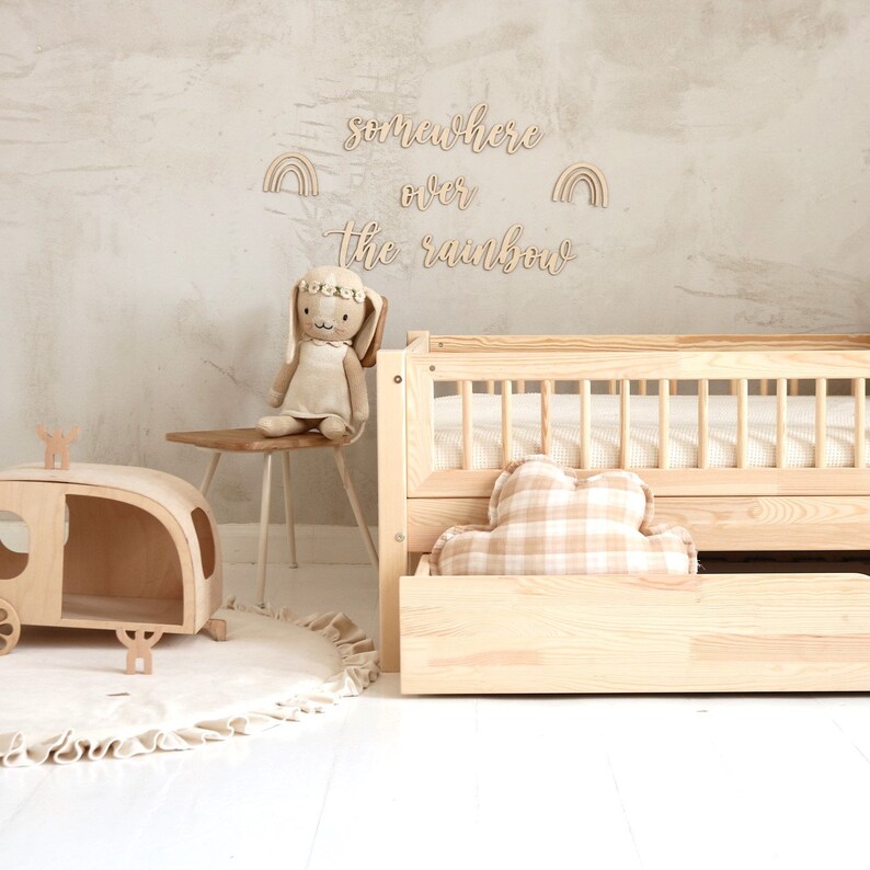 K&ouml;nnte beinhalten: Ein Kinderbett aus Holz mit einem wei&szlig;-braunen karierten Kissen und einem h&ouml;lzernen Spielzeug-Wohnwagen. Die Wand hinter dem Bett hat ein Holzschild mit der Aufschrift "somewhere over the rainbow" mit zwei Regenbogen-Ausschnitten.