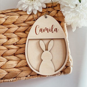 May include: A wooden Easter egg ornament with the name "Camila" engraved on it. The egg has a cutout shape of a bunny rabbit inside.