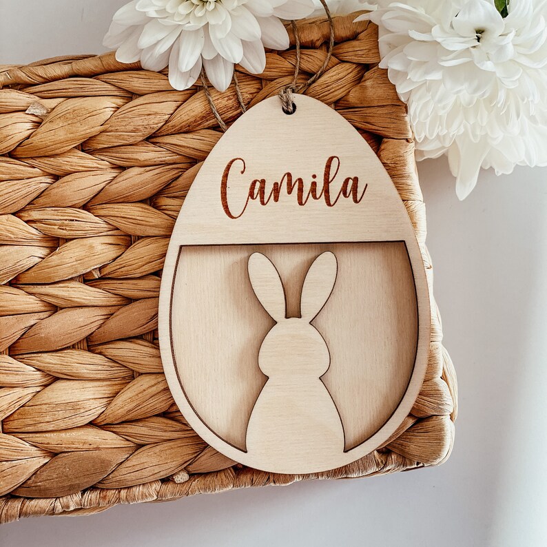 May include: A wooden Easter egg ornament with the name "Camila" engraved on it. The egg has a cutout shape of a bunny rabbit inside.