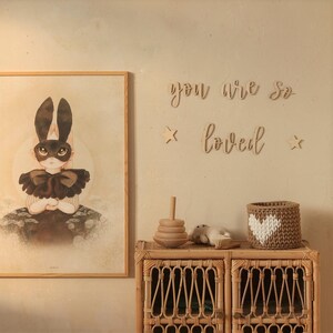 May include: A wooden sign with the words "you are so loved" in cursive script, with two small wooden stars on either side. The sign is hanging on a light beige wall above a wicker cabinet with a wooden toy stacking ring and a crocheted heart-shaped basket.