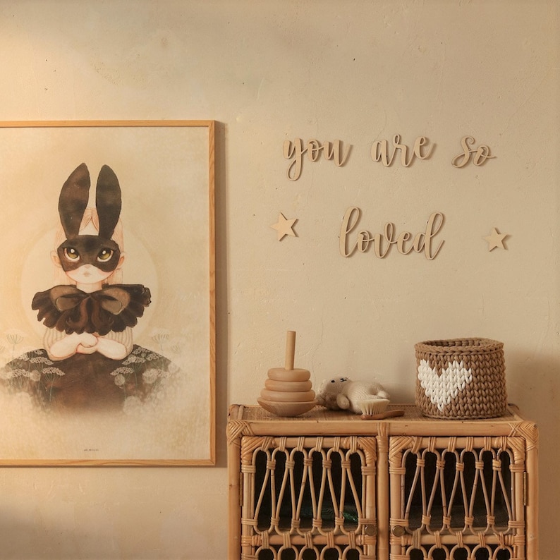 May include: A wooden sign with the words "you are so loved" in cursive script, with two small wooden stars on either side. The sign is hanging on a light beige wall above a wicker cabinet with a wooden toy stacking ring and a crocheted heart-shaped basket.