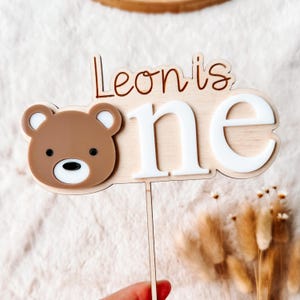Puede incluir: Un adorno para tarta de madera con el texto "Leon is one". El adorno presenta una cabeza de oso marr&oacute;n con detalles blancos y la palabra "one" en blanco. El dise&ntilde;o est&aacute; sobre una base de madera clara, perfecto para una celebraci&oacute;n de primer cumplea&ntilde;os.