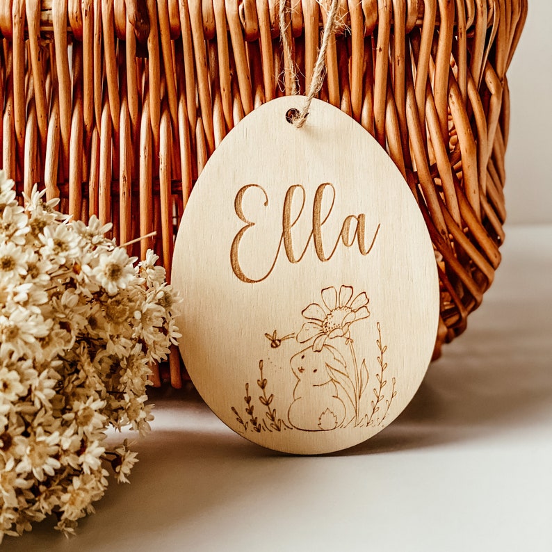 Op de afbeelding: Een houten eivormige hanger met de naam "Ella" erop gegraveerd. De hanger heeft een klein konijn dat naast een bloem zit.