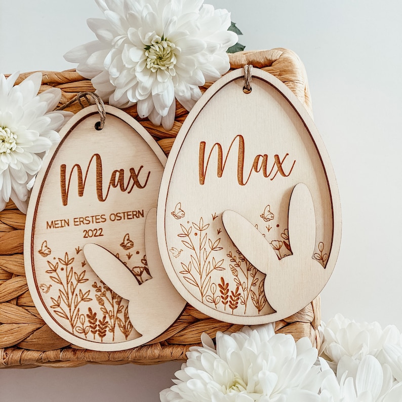 May include: Two wooden Easter egg ornaments with the name Max engraved on them. One egg has the text "Mein erstes Ostern 2022" engraved on it. Both eggs have a bunny rabbit cutout and floral designs.