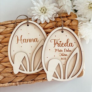 May include: Two wooden Easter egg ornaments with bunny ear cutouts. One ornament says "Hanna" and the other says "Frieda: Mein Erstes Ostern 2022".