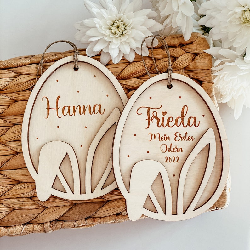 May include: Two wooden Easter egg ornaments with bunny ear cutouts. One ornament says "Hanna" and the other says "Frieda: Mein Erstes Ostern 2022".