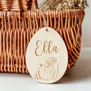 Op de afbeelding: Een houten paasei-versiering met de naam "Ella" erop gegraveerd. De versiering heeft een kleine konijnenillustratie met een bloem en een bij.