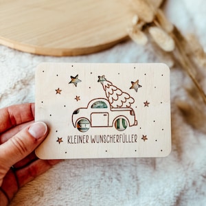 K&ouml;nnte beinhalten: Eine Holzkarte mit einem Design eines Autos, das einen Weihnachtsbaum transportiert. Die Karte hat ausgeschnittene Sterne und den Text "KLEINER WUNSCHERF&Uuml;LLER". Die Karte wird von einer Hand gehalten.