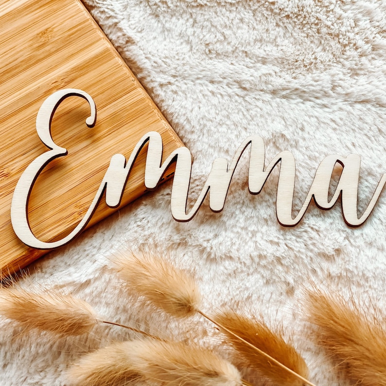 Puede incluir: Letrero de nombre de madera que deletrea "Emma" en una fuente cursiva. El letrero est&aacute; sobre una superficie peluda de color marr&oacute;n claro con plantas secas parecidas a la hierba.