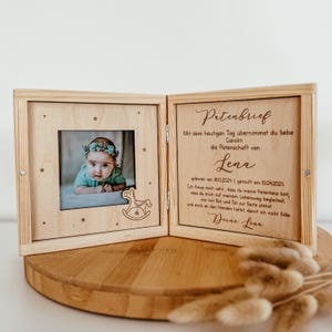 May include: A wooden box with a hinged lid, containing a photo of a baby and a handwritten letter. The letter is addressed to "Carolin" and states that she is taking on the role of godparent for "Lena", who was born on 18/10/2024 and baptized on 15/04/2025. The letter is signed "Deine Lena".