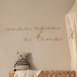 May include: Wooden wall decor with the words "once upon a time" in a cursive font. The decor is displayed on a beige wall with a stuffed animal and a canopy bed in the background.