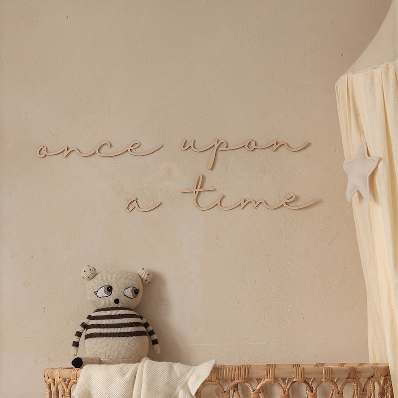 May include: Wooden wall decor with the words "once upon a time" in a cursive font. The decor is displayed on a beige wall with a stuffed animal and a canopy bed in the background.