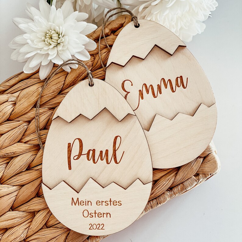May include: Two wooden Easter egg ornaments with the names "Emma" and "Paul" engraved on them. The ornament for Paul also has the text "Mein erstes Ostern 2022" engraved on it.
