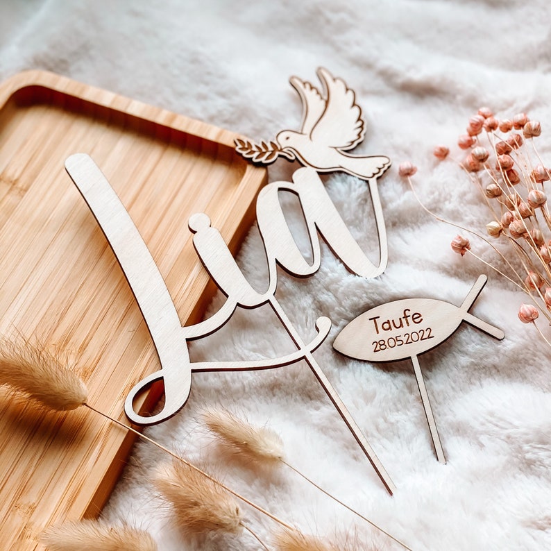 May include: Wooden cake topper with the name "Lia" in cursive script, a dove with an olive branch, and a fish with the text "Taufe 28.05.2022".